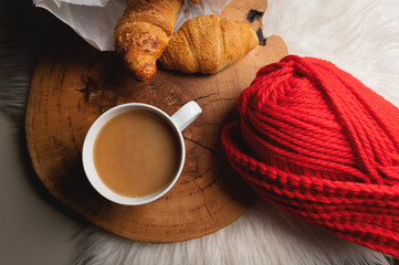 Atmospheric still life white mug of coffee next to fresh croissants in a paper bag next to red yarn. Home comfort in soft focus