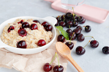 Oatmeal porridge with fresh black cherry in a white bowl, wooden spoon and cherries with leaves on white table. Healthy eating concept. Close-up. Selective focus. Blurred background.