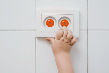 A small child's hand touches an electrical outlet with child safety plugs. The child inserts his...