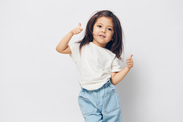 happy, joyful, little girl stands in a white t-shirt and shows thumbs up joyfully looking at the camera. Horizontal photo on a light background with empty space for an advertising layout insert