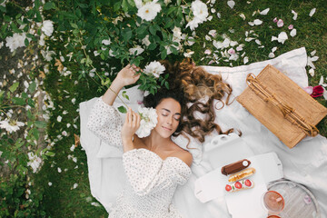 Young beautiful woman with long curly hair having picnic with champagne and fresh eclairs in...
