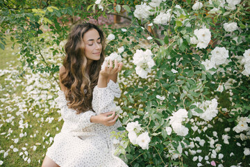 Young beautiful woman with long curly hair wearing white dress relaxing in blooming rose garden.