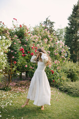 Young beautiful woman with long curly hair wearing white dress relaxing in blooming rose garden.