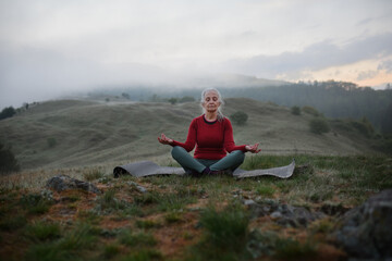 Naklejka na ściany i meble Senior woman doing breathing exercise in nature on early morning with fog and mountains in background.