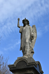 Worn stone sculpture of an angel pointing skyward while looking down, with blue sky and light clouds in the background
