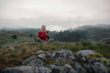 Naklejka na ściany i meble Senior woman taking selfie when doing breathing exercise in nature on early morning with fog and mountains in background.