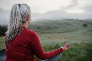 Rear view of senior woman doing breathing exercise in nature on early morning with fog and...