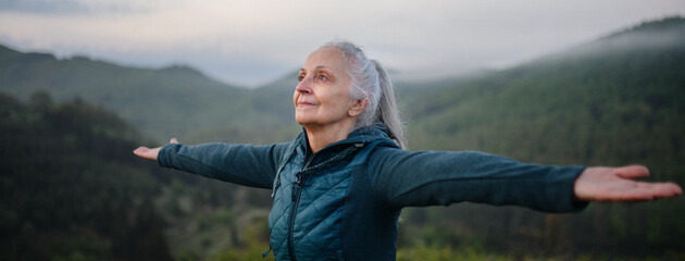 Senior woman doing breathing exercise in nature on early morning with fog and mountains in background. - obrazy, fototapety, plakaty