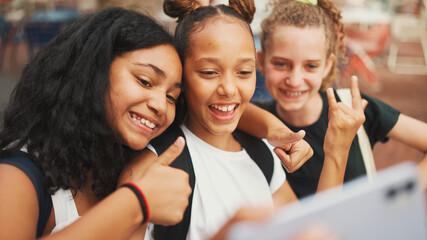 Three girls friends pre-teenage standing on the street smiling, hugging each other making faces for the cell phone camera, selfie. Teenagers on the outdoors in urban cityscape background