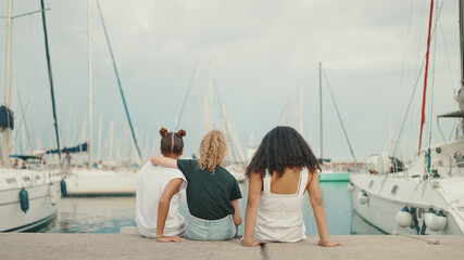 Laughing three girls friends pre-teenage sitting on the waterfront against ships and yachts background. Teenagers singing and clapping on the outdoors in seascape background