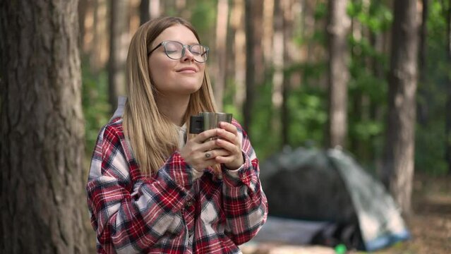 Satisfied Caucasian young woman drinking tea from thermos smiling looking around standing in forest outdoors. Medium shot portrait of happy relaxed beautiful traveler enjoying leisure camping