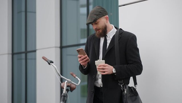 A handsome man in a suit looks at the phone drinking coffee near business center. Man wearing a cap and shoulder bag, standing near black old-fashioned bicycle. High quality 4k footage