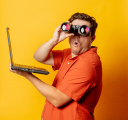 Guy in orange shirt watch in binoculars and laptop computer on yellow background