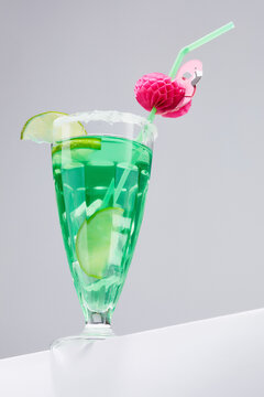 Close-up shot of a glass of green cocktail with lime and ice. The cocktail is decorated with a green corrugated curved straw with a pink flamingo decor. The glass stands on a gray background. 