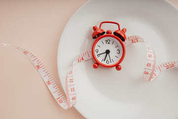 Time to lose weight concept. Red alarm clock with white plate and measuring tape close-up
