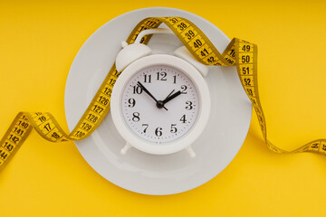 Diet concept. Alarm clock on a white plate with measuring tape