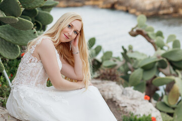 Fototapeta na wymiar Portrait of young smiling attractive woman bride sitting on rock near green huge leaves of opuntia cactus. Wedding.