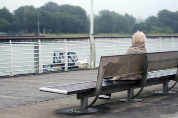 Lonely old lady gazes at the sea.
She lady sitting on a bench thinking about life.
Emotions and memories on a cloudy day.