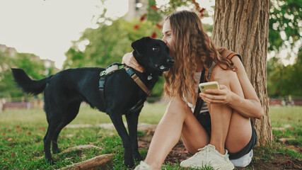 Cute girl with black dog is resting under tree on the lawn. Happy dog rejoices being on walk in the outdoor. Animal training outdoors.