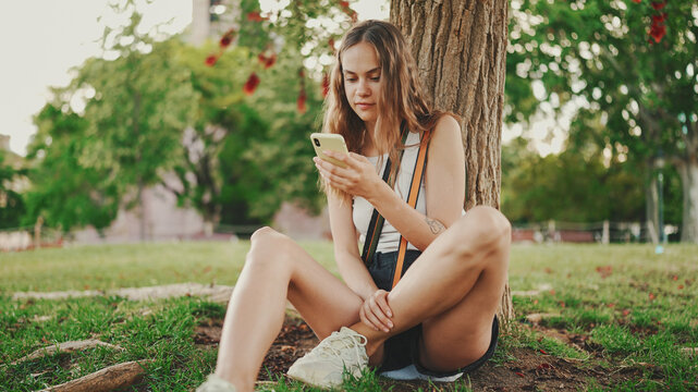 Beautiful girl with long wavy hair wearing in white top uses mobile phone while sitting on the green grass under tree in the park. Girl flip through the pictures on your phone with fingers