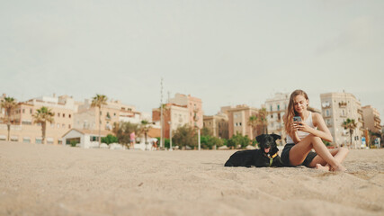 Fototapeta na wymiar Lunch time. cute girl is sitting on the sand on the beach with her pet testing sandwich. The girl treats her dog with sandwich. Lunch on the beach on modern city background