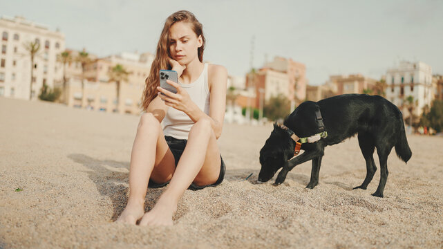 Cute girl with mobile phone in her hands sits on the sand on the beach with black dog on modern building background. Beautiful girl is watching videos and photos on her smartphone