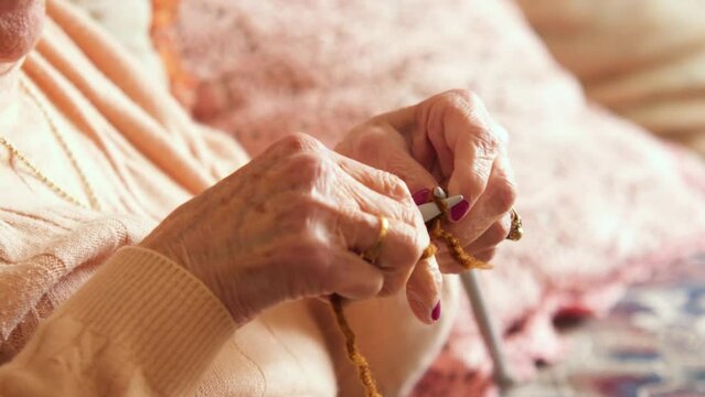 An old woman knitting in her living room, receiving sunlight, with a pink pillow behind. Her jaw is shaking due to concentration. Parkinson disease