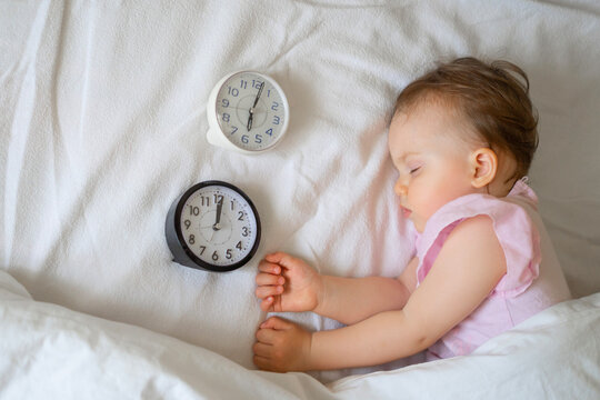 A Little Girl Sleeps In A White Bed With A Clock. Sleep Schedule For A Child. Alarm Feeding