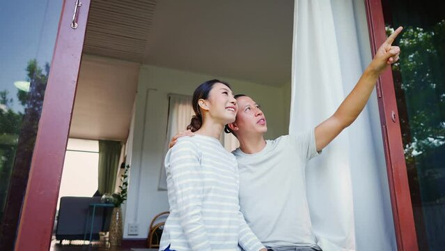 Portrait of young adult Asian couple looking up while a man is pointing with a finger in home interior background. 30s mature successful husband and wife - 4K Slow Motion Footage