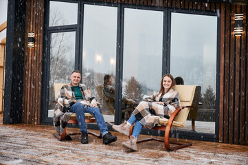 Obraz na płótnie Canvas Cheerful man and woman wrapped in blanket sitting in chairs outside scandinavian house barnhouse. Happy couple resting together outdoors near building with panoramic window under winter snow.