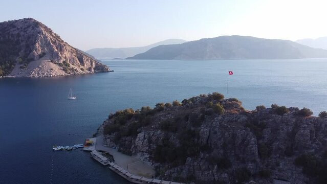 Rock in sea bay with sand beach and turkish flag on top in small touristic village Turunc. Landscape with moored pleasure yacht. Drone view