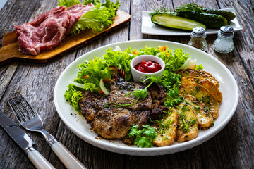 Roasted pork loin and fried potatoes with green vegetables on wooden table
