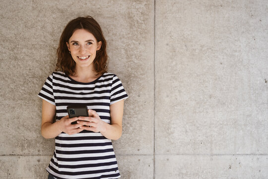 Young happy woman with cellphone stands in front of modern brown wall and looks to camera