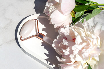Rose quartz face roller for beauty facial massage therapy. Skin care tool for massaging morning routine. Luxury product on white marble background with peony flowers. Top view.