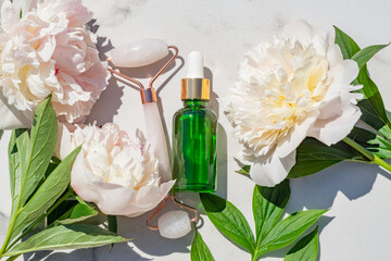 Face massage roller with natural essential oil in green pipette bottle on marble background with peonies. Flat lay, top view. Facial skin care, body treatment concept.