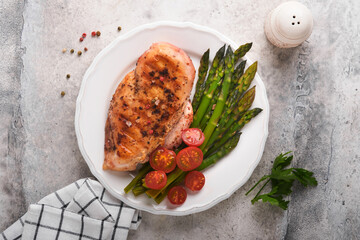Chicken breast grilled with vegetables. Grilled chicken steak, asparagus and cherry tomatoes in black plate on light gray concrete table background. Healthy diet lunch. Barbecue steak fried. Top view.
