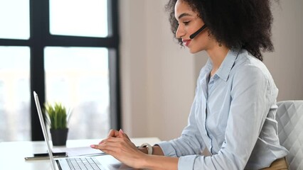 Side view of an intelligent young mixed-race woman with Afro hairstyle in a headset in the office, takes a call and smiling, working with a laptop in the sales department or as a call center operator