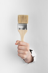 child hand through hole in white paper with torn edges holding a paint brush, copy space