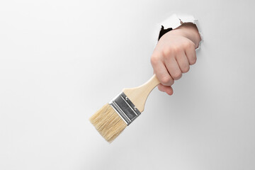 child hand through hole in white paper with torn edges holding a paint brush, copy space