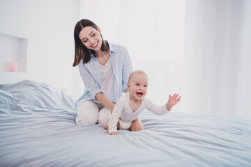 Fototapeta na wymiar Photo of charming funny small baby mom wear blue shirt holding crawling bed smiling indoors home room