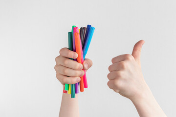 child hand holding many colorful felt-tip pens and showing thumbs up above a white and gray background, copy space - obrazy, fototapety, plakaty
