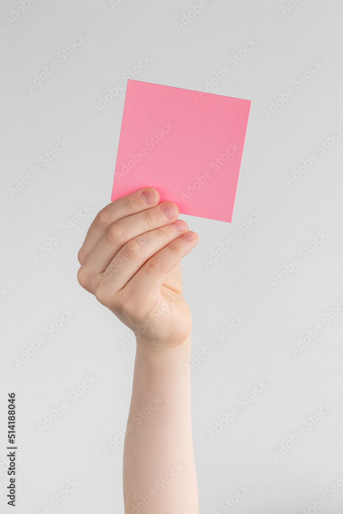 Wall mural child hand holding a square pink blank reminder or paper notes above a white and gray background, copy space