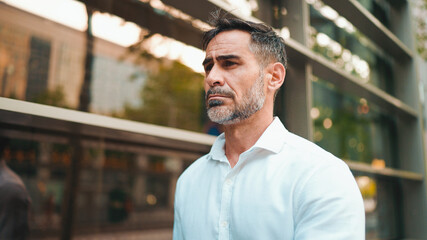 Fototapeta premium Mature businessman with neat beard wearing white shirt on his way to the office in the financial district in the city. Successful man Looks at the upper floors of modern buildings