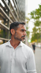 Mature businessman with neat beard wearing white shirt on his way to the office in the financial district in the city. Successful man Looks at the upper floors of modern buildings