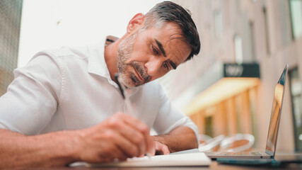 Mature businessman with neat beard wearing white shirt writing down notes in paper notepad, sitting...