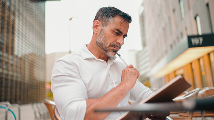 Fototapeta premium Mature businessman with neat beard wearing white shirt writing down notes in paper notepad, sitting at table. making appointments daily tasks in personal organizer