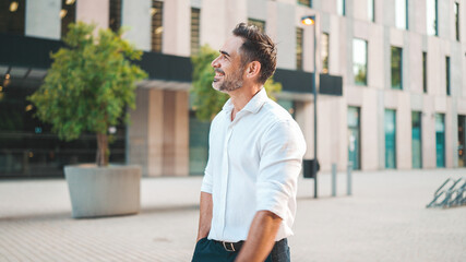 Mature businessman with neat beard wearing white shirt on his way to the office in the financial district in the city. Successful man Looks at the upper floors of modern buildings