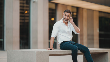 Mature businessman with neat beard wearing white shirt uses mobile phone sits on bench in the financial district in the city. Successful man talking on smartphone.