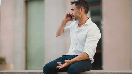 Close-up mature businessman with neat beard uses mobile phone sits on bench in the financial...