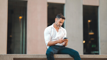 Mature businessman with neat beard uses mobile phone sits on bench in the financial district in the city. Successful man scrolls through information on smartphone and smiles